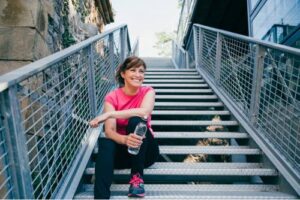 A woman wearing a pink shirt is sitting on stairs, smiling, and holding a water bottle after a workout.