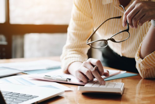 Close-up of a person using a calculator and holding up glasses while working on a desk with a computer and finance files on it.