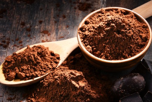 A close-up of cocoa powder in a wooden bowl and spoon on a rustic wooden surface, with chocolate pieces nearby.