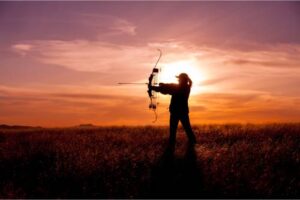 A woman with a bow forms a silhouette in front of a setting sun. She is in an open field pointing her arrow.