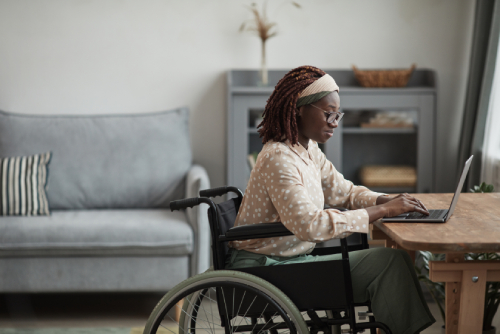 An African-American woman sits in her wheelchair at a kitchen table. She is typing on a laptop in front of her.
