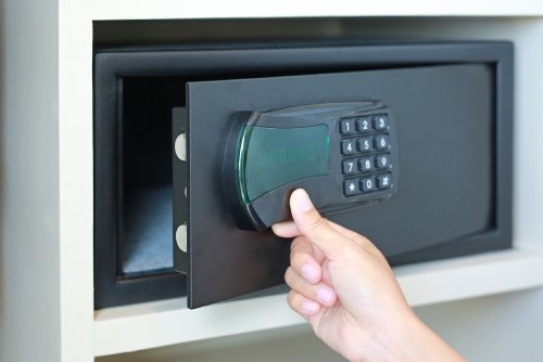 A person's hand opening a black safe located on a rectangular shelf. The front of the safe has a keypad.