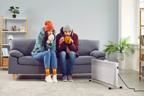 A man and woman are sitting on a couch indoors, huddling together, looking cold and wearing insulating clothes.