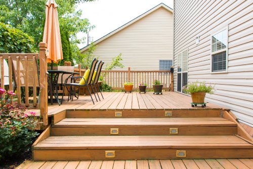 A large, multi-level wooden deck with stairs, railings, outdoor furniture, and plants sits in the backyard of a home.