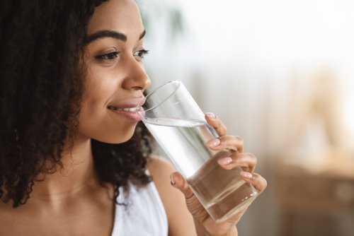 A woman in a white shirt smiles softly as she holds a glass of water to her lips in a sunny room.