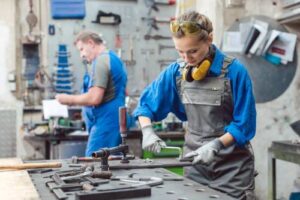 A woman wearing a blue shirt and a hard hat works alongside a man in an industrial job in a workshop.