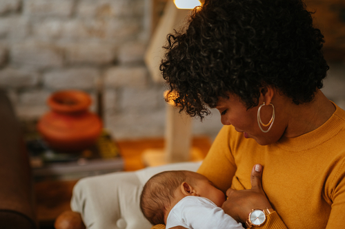 A mother wearing a yellow shirt and gold watch is breastfeeding her child. The background is blurry.
