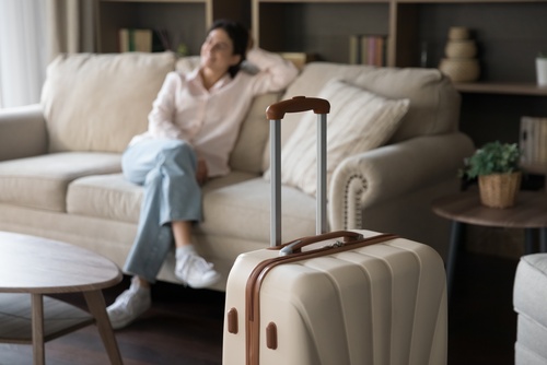 A blurred woman sitting on a couch with her legs crossed. A beige and brown suitcase is near the camera.