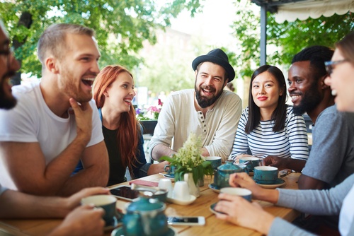 A group of friends sit at a table at a cafe, some smiling and others laughing. Some hold a coffee cup in their hands.