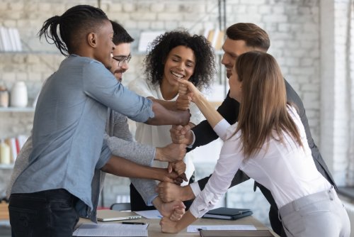 A group of people stand around a table and create a tower with their hands. They are all smiling while they're doing it.